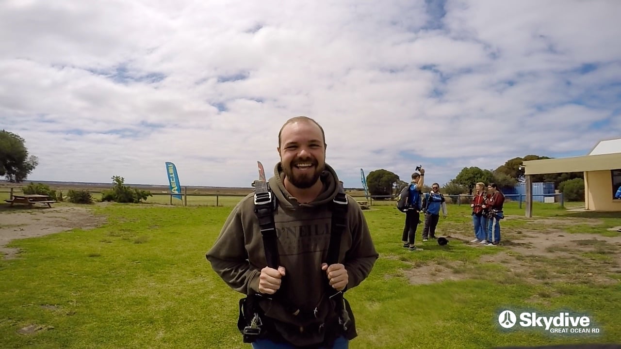 A smiling, bearded young man in a skydiving harness on the grass at a coastal skydive center, others waiting behind him.