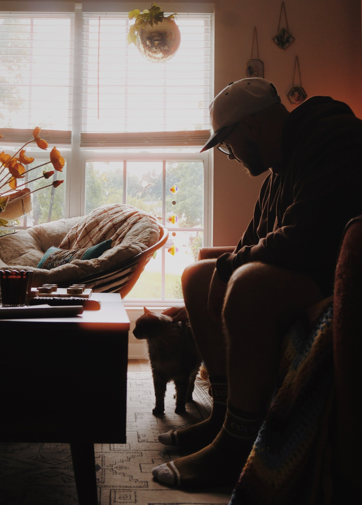 Ian in a cap sitting in the shadowy front room, a cat at his feet, sunlight through the window behind.