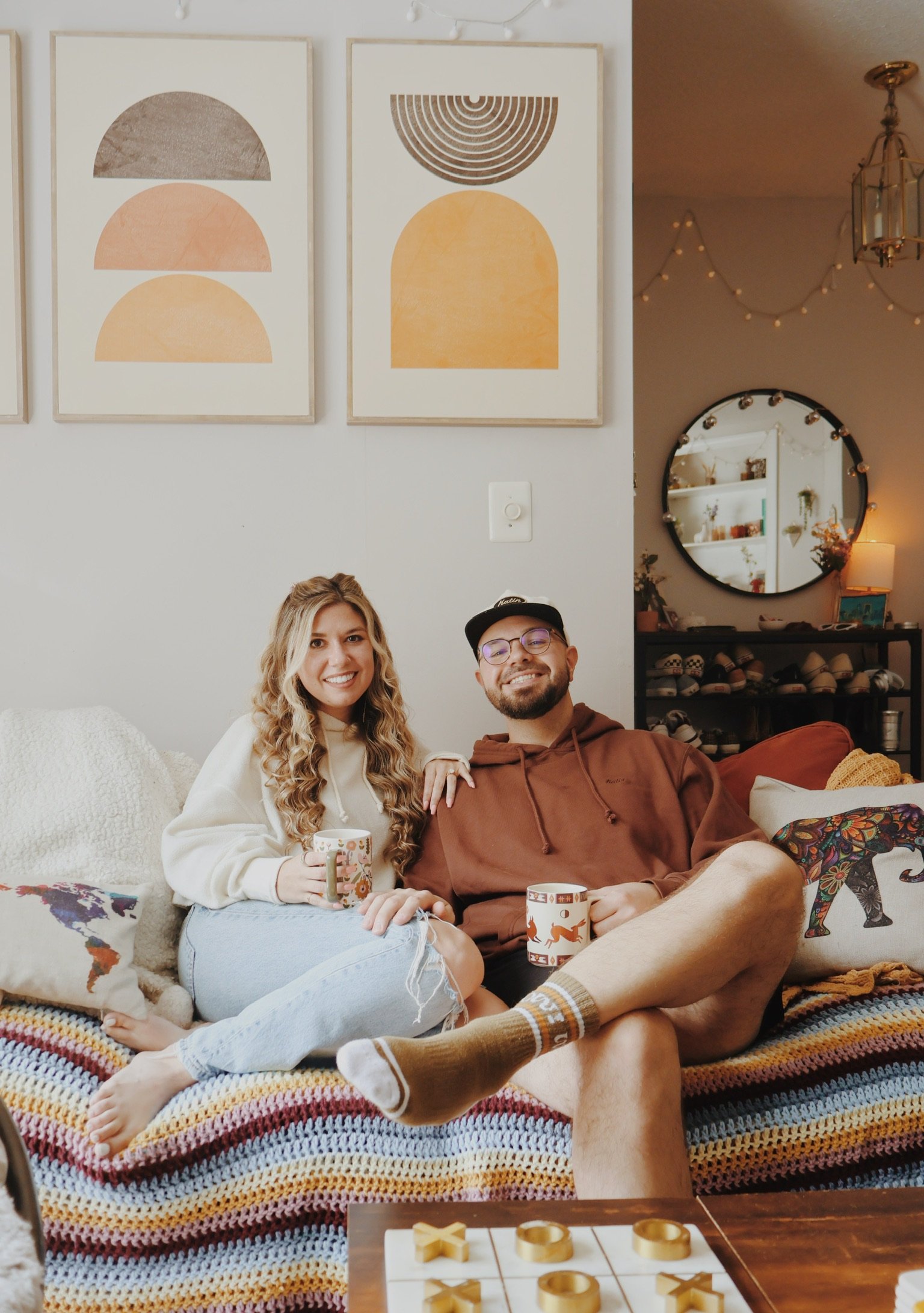 Nicole and Ian with their feet up on the striped couch, mugs in hand, under two arched art prints.