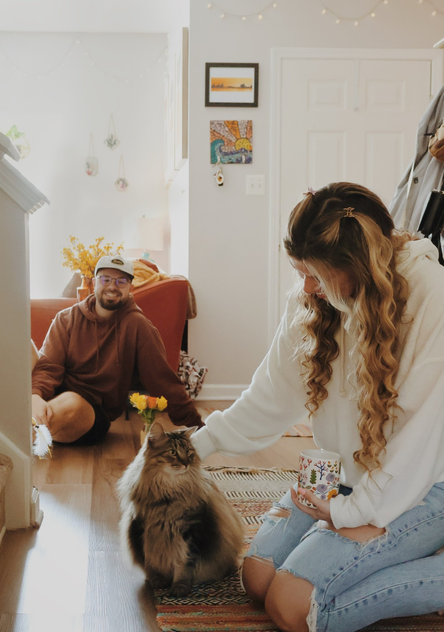 Nicole kneeling to stroke a big long-haired cat on the wooden floor while Ian watches from the chair.