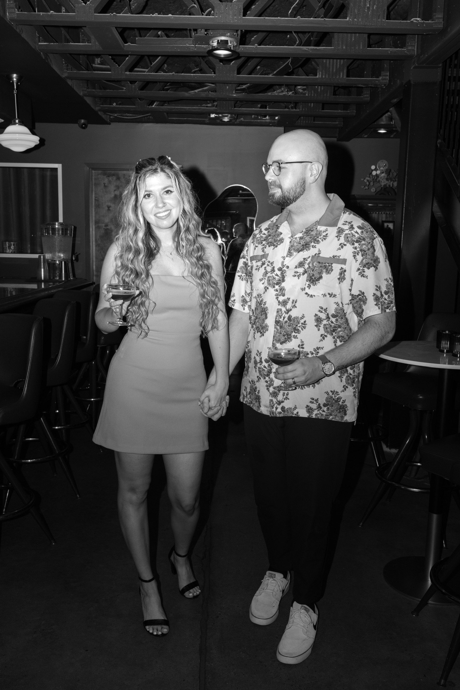 In black and white: Nicole and Ian walking hand in hand across the dark bar, drinks in hand.