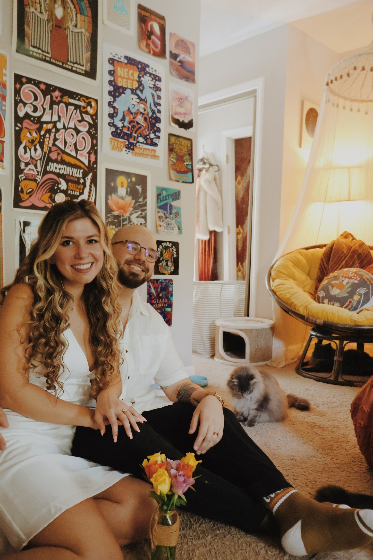 Nicole and Ian in white sitting on the floor with a jar of flowers, a cat walking past behind them.
