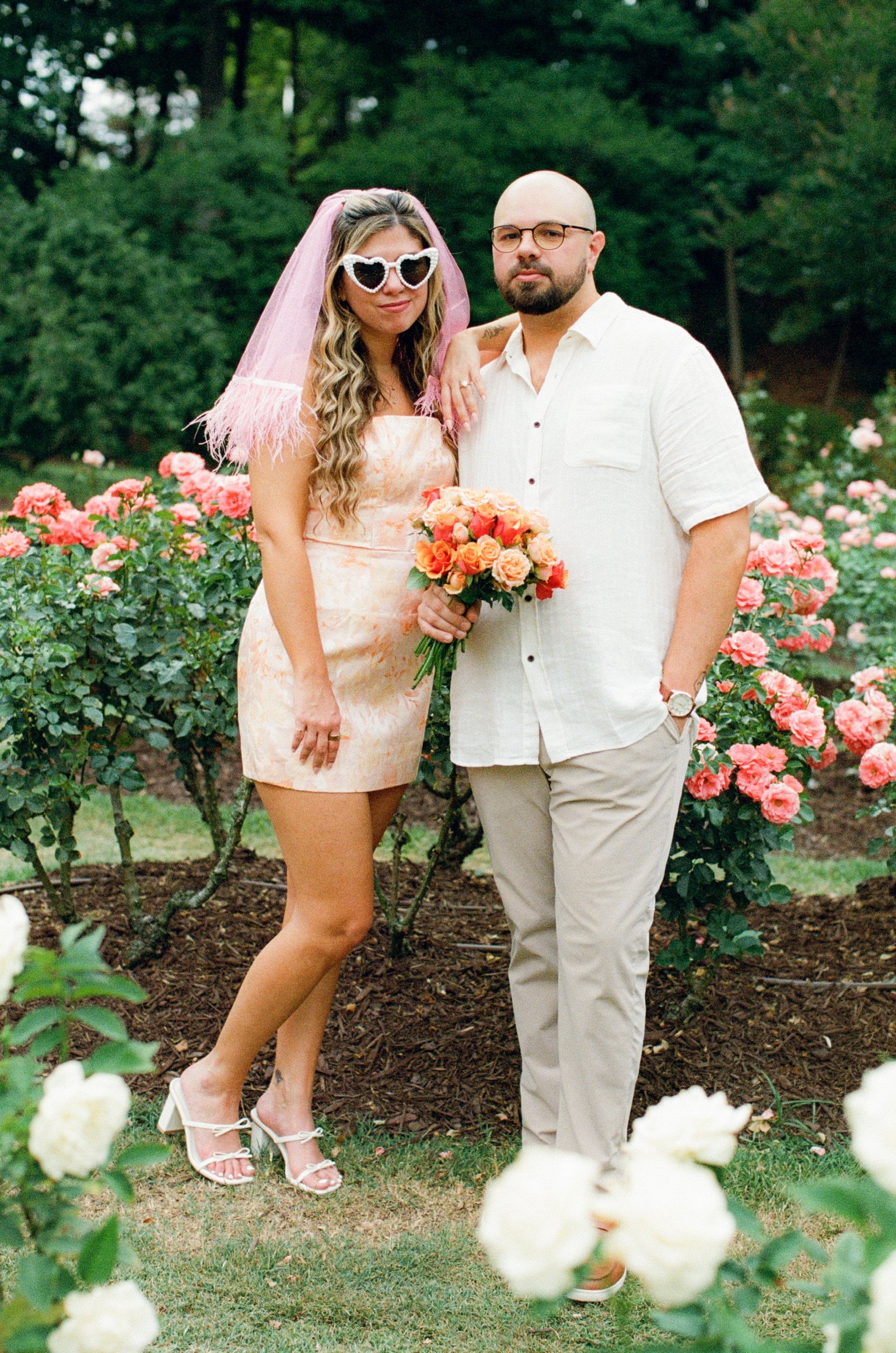 Nicole in heart-shaped sunglasses and a pink feathered veil, holding orange roses beside Ian.