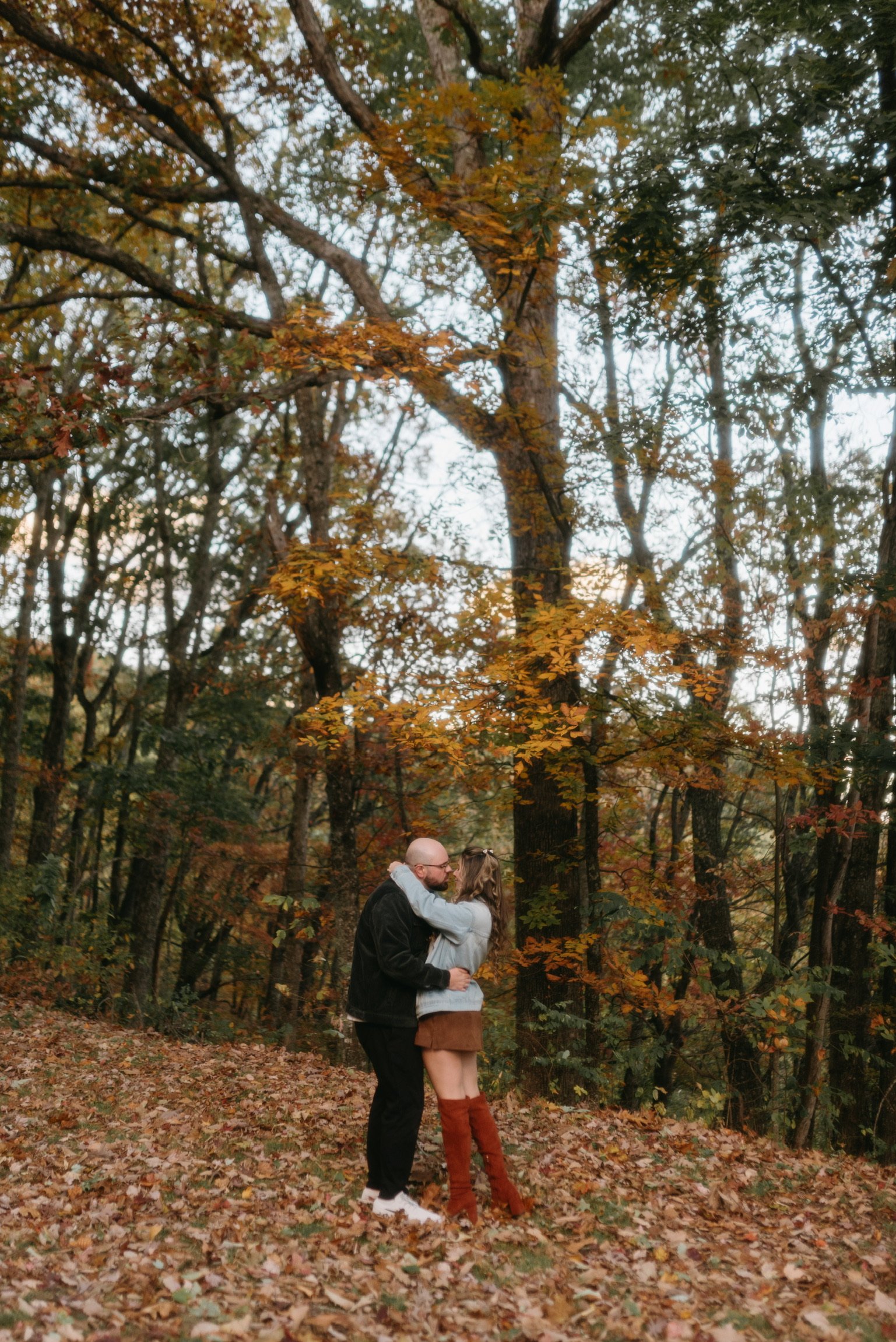 Nicole and Ian holding each other on a carpet of leaves beneath tall autumn trees.