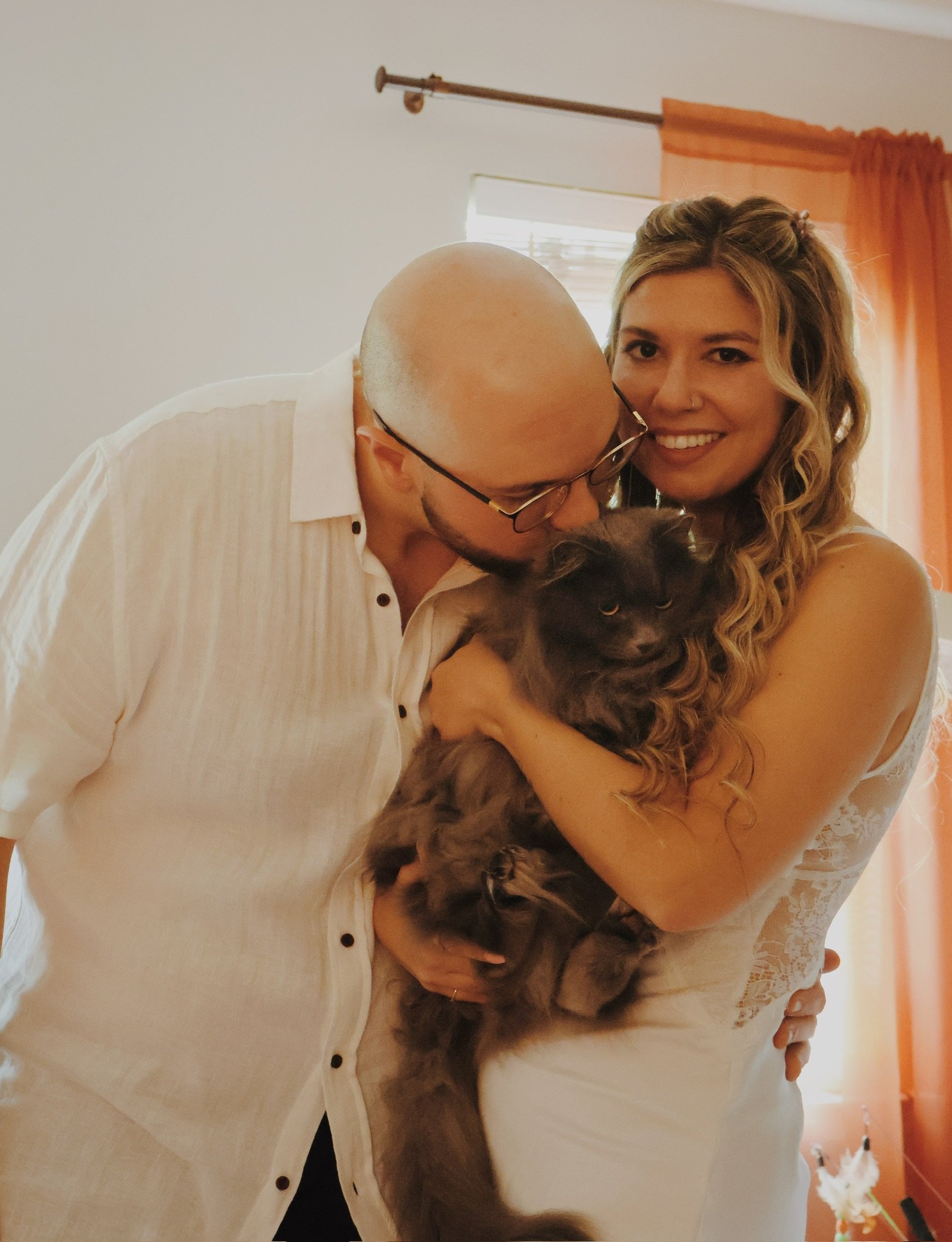 Ian and Nicole in white, holding a fluffy cat between them and smiling.
