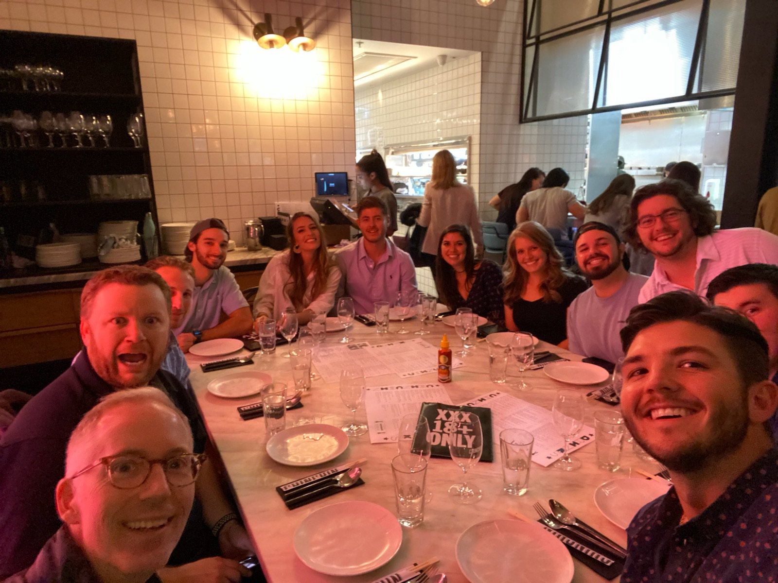 A long table of friends at dinner in a tiled restaurant, leaning in for a selfie.
