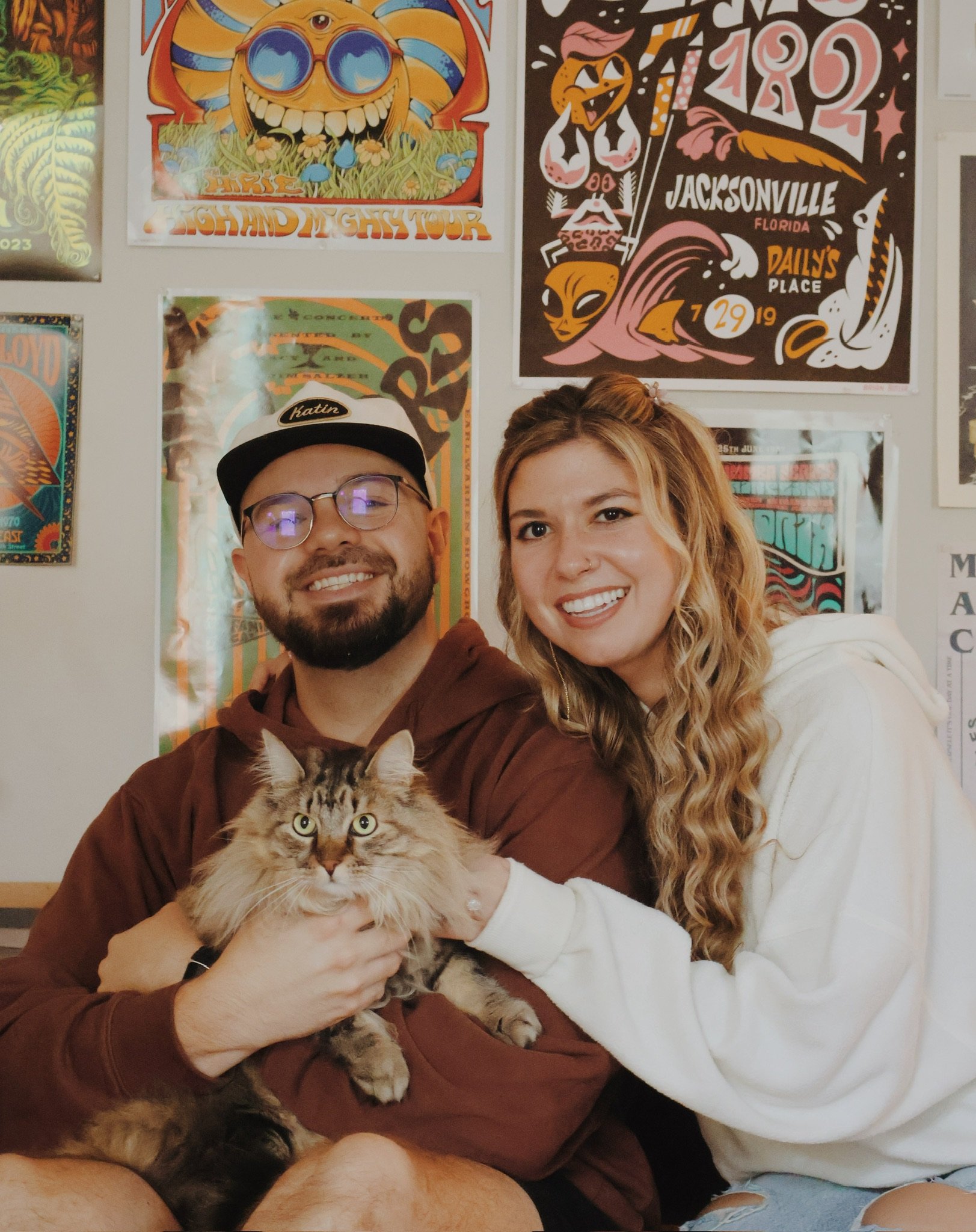Ian holding a long-haired tabby cat as Nicole hugs his arm, gig posters behind them.