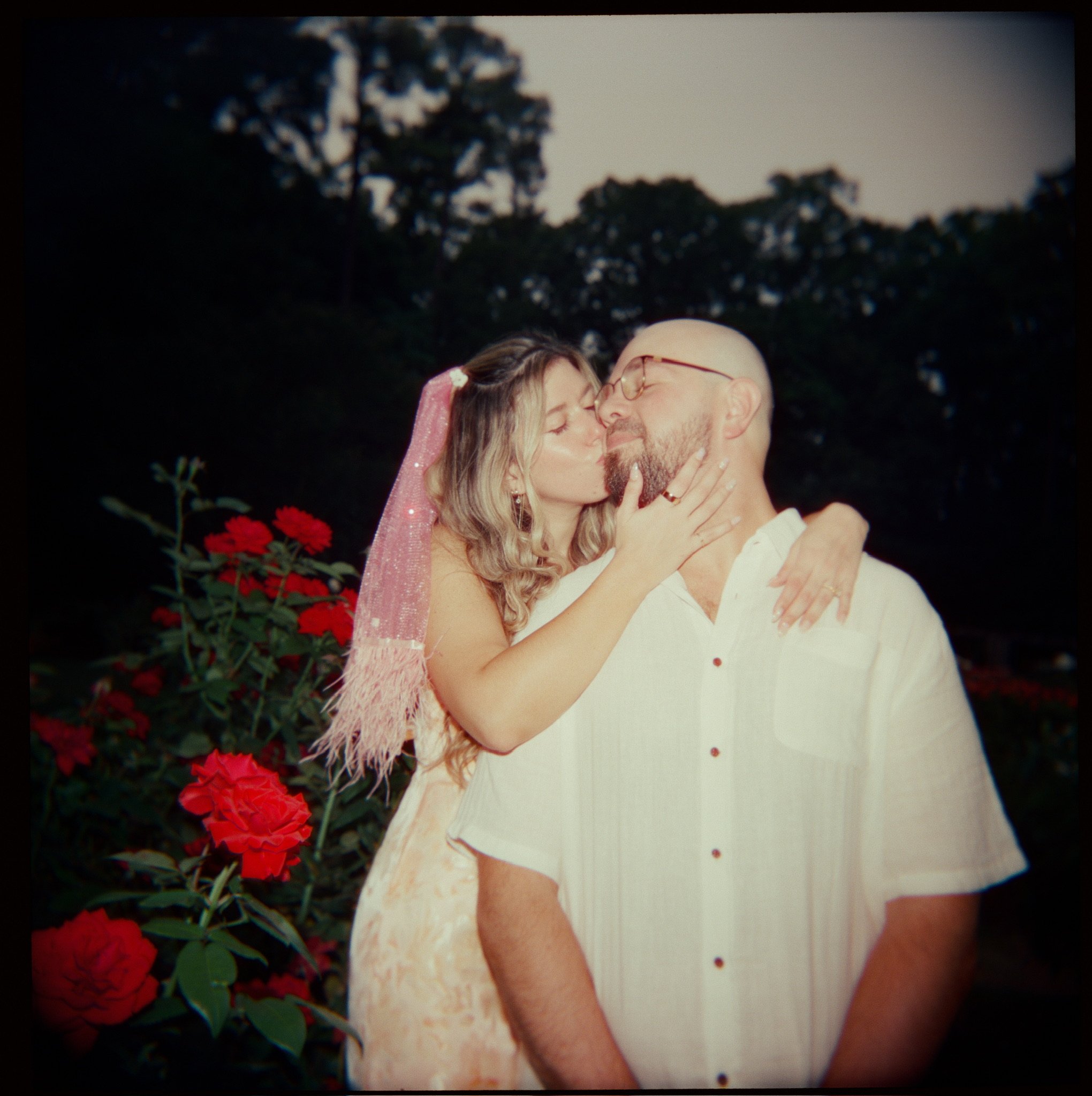 Nicole kissing Ian’s cheek in front of deep red roses as evening falls.