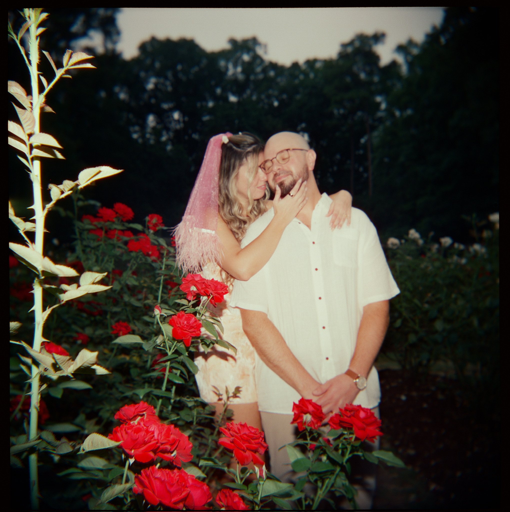 Nicole with her arms around Ian’s neck among red roses, a young stem in the foreground.