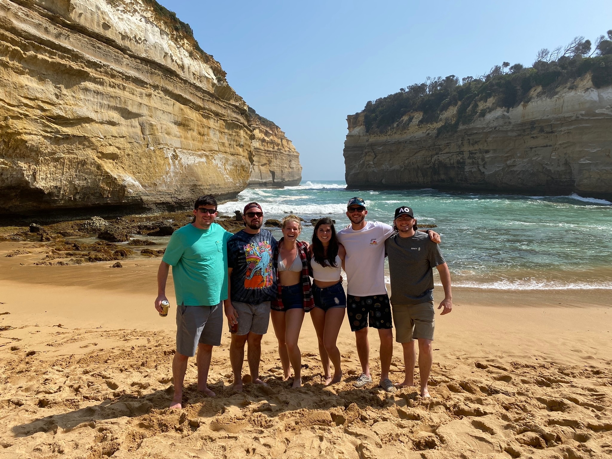 Six friends on a beach between two tall sandstone cliffs, waves rolling in behind them.
