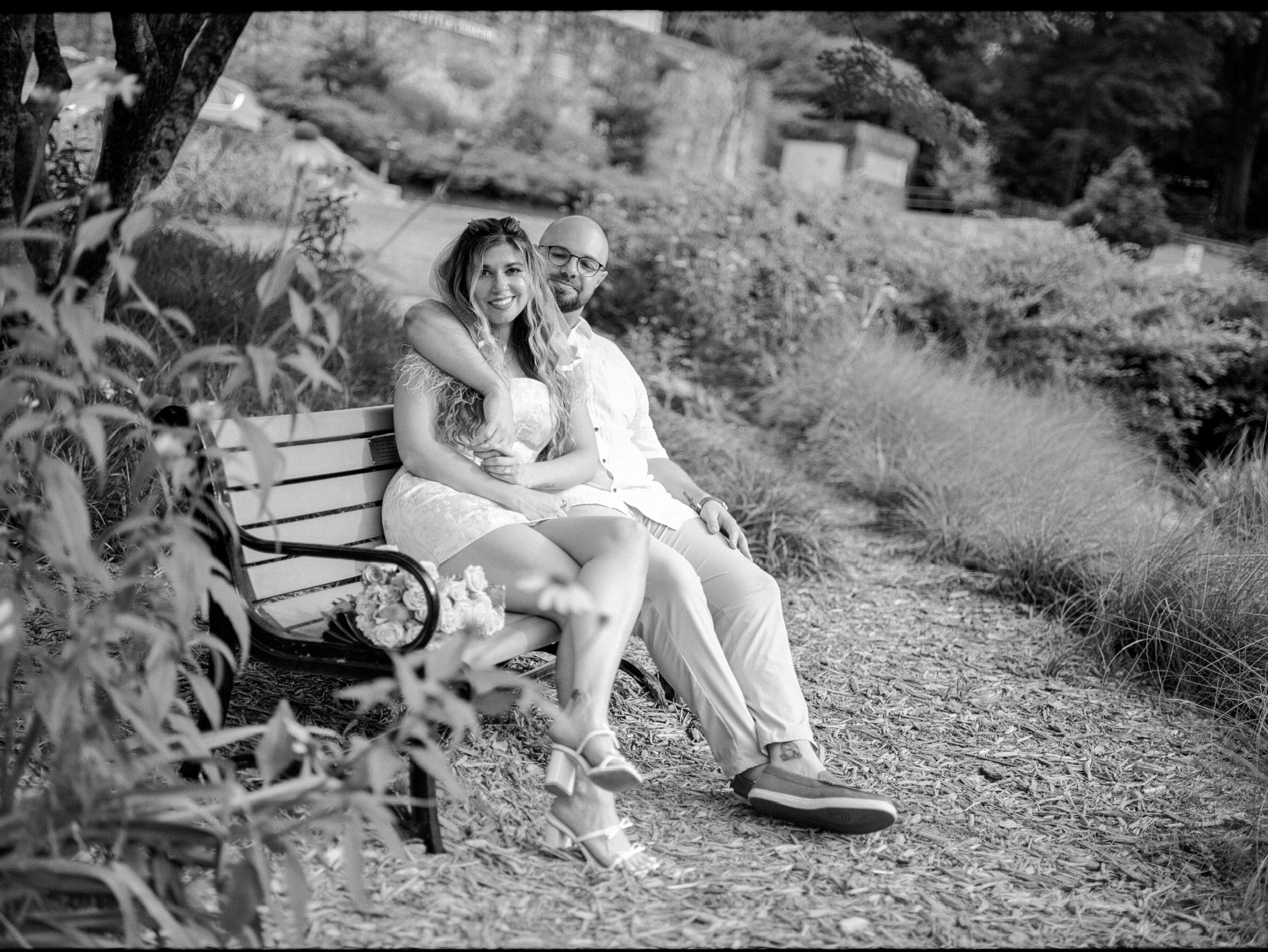 In black and white: Nicole and Ian sitting close on a park bench beside the path.