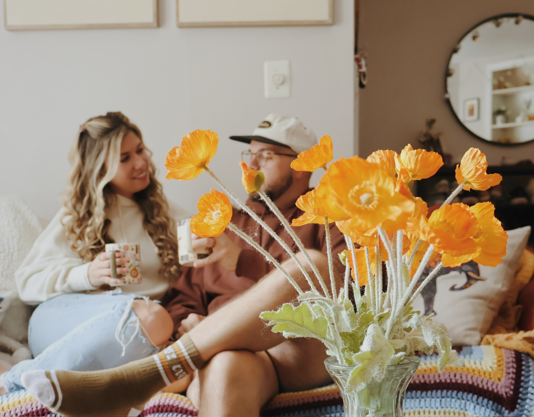 Nicole and Ian on the couch with mugs, a vase of orange poppies in the foreground.