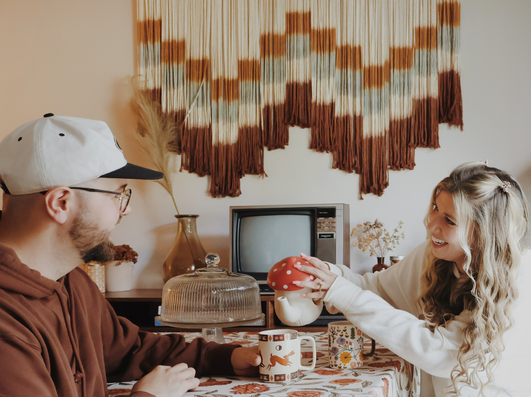 Nicole setting a toadstool-shaped cookie jar on the table as Ian looks on, a macramé wall hanging behind.