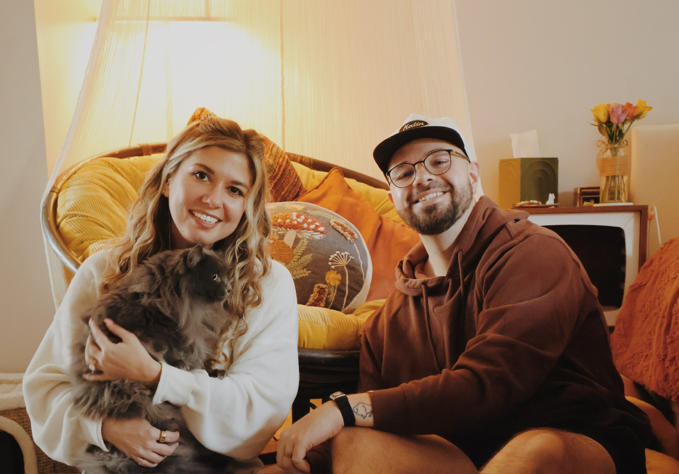 Nicole holding a gray cat in a big papasan chair, Ian smiling beside her in the lamplight.