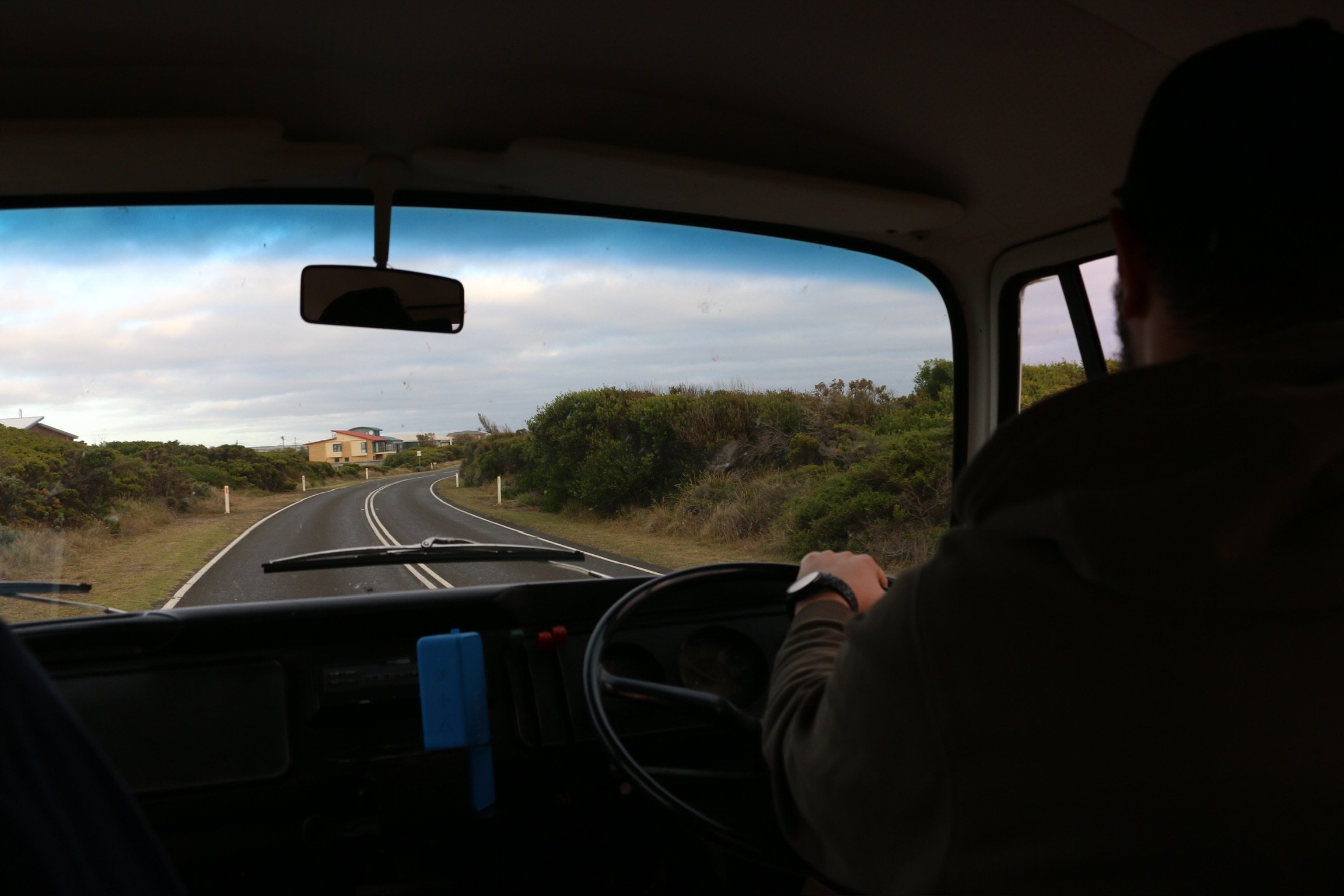 From inside the kombi: the driver’s arm on the wide steering wheel as the coast road curves ahead.