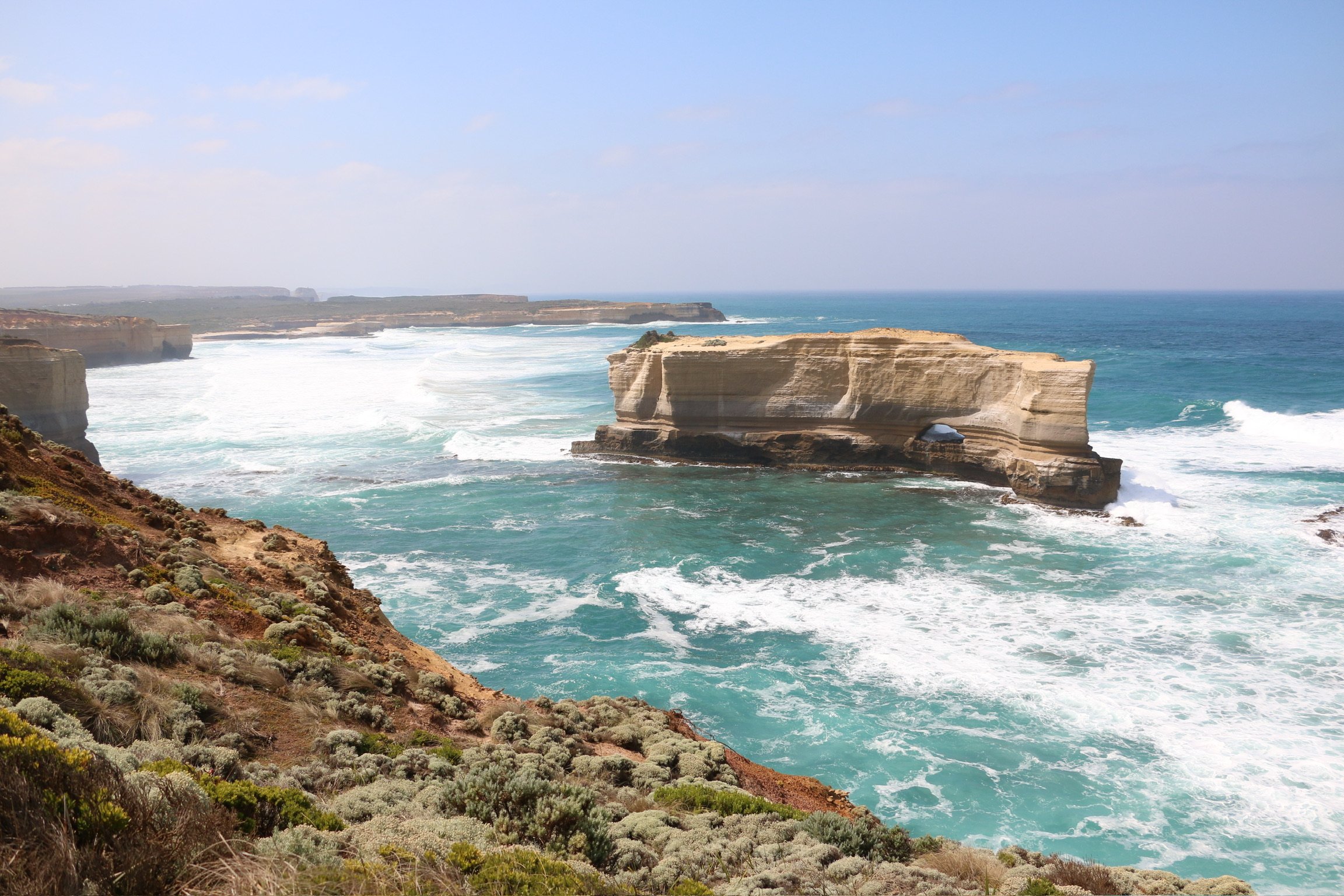 Turquoise surf breaking around a flat-topped sandstone sea stack off the coast.